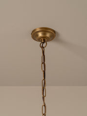 A pleated conical pendant light shade in a warm cream or off-white fabric, illuminated with a soft warm glow. It hangs from a dark brass chain and ceiling canopy against a plain, warm beige wall.