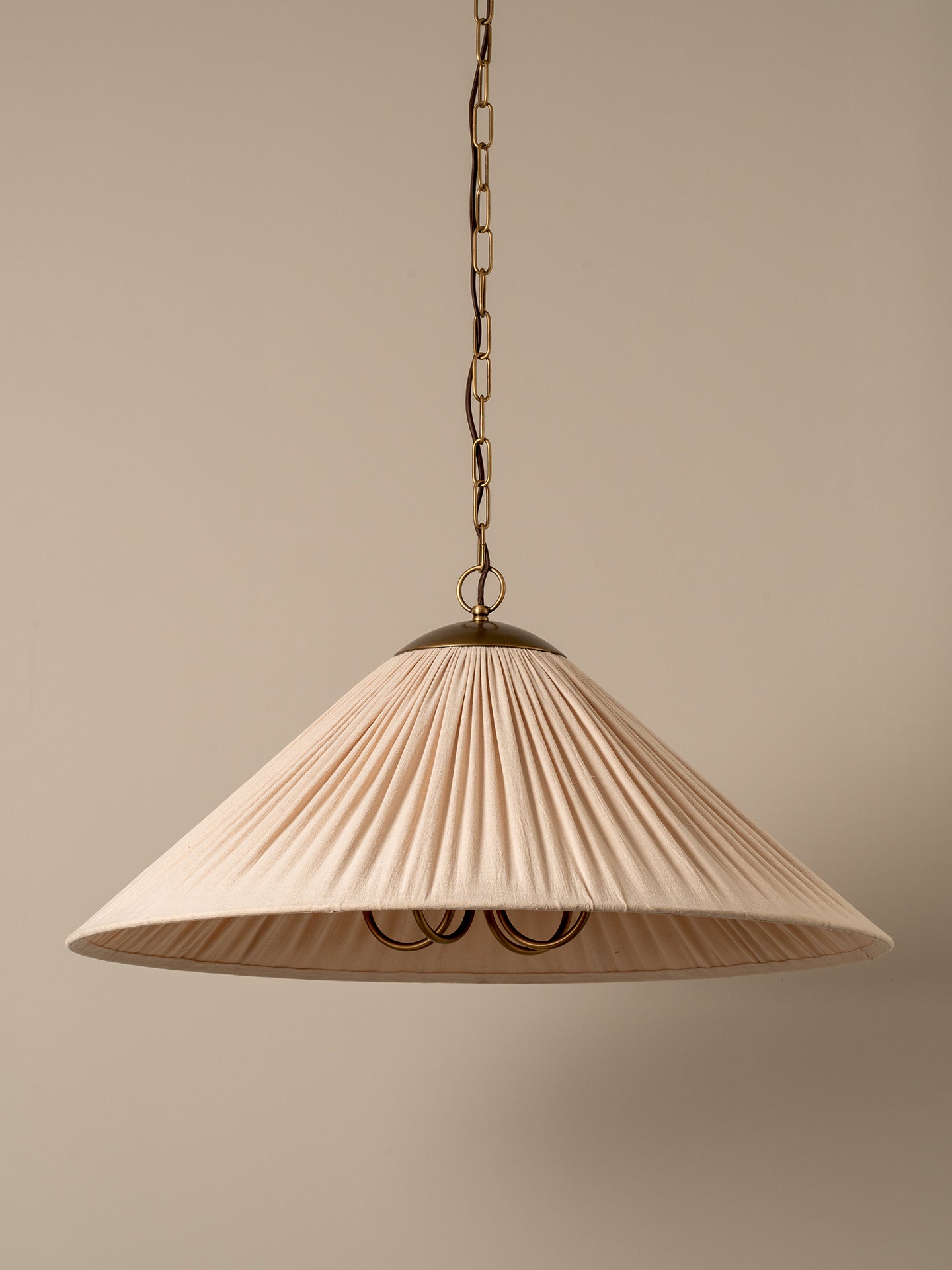 A pleated conical pendant light shade in a warm cream or off-white fabric, illuminated with a soft warm glow. It hangs from a dark brass chain and ceiling canopy against a plain, warm beige wall.