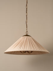 A pleated conical pendant light shade in a warm cream or off-white fabric, illuminated with a soft warm glow. It hangs from a dark brass chain and ceiling canopy against a plain, warm beige wall.
