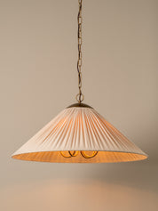 A pleated fabric pendant lampshade in a warm cream or off-white color, suspended from a brass chain with a warm light glowing from within, set against a solid neutral beige background.