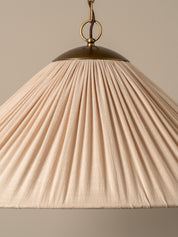A pleated conical pendant light shade in a warm cream or off-white fabric, illuminated with a soft warm glow. It hangs from a dark brass chain and ceiling canopy against a plain, warm beige wall.