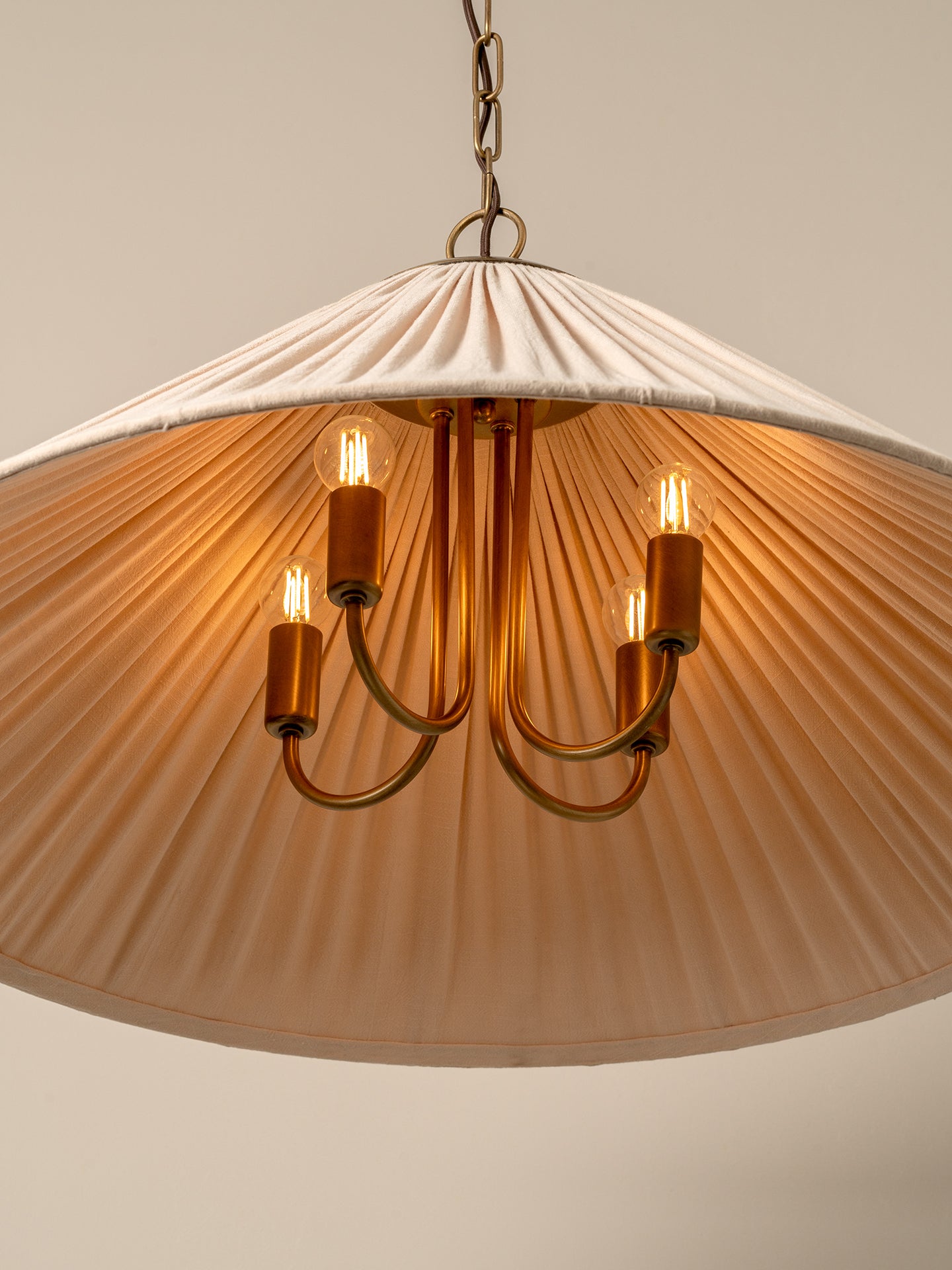 A pleated conical pendant light shade in a warm cream or off-white fabric, illuminated with a soft warm glow. It hangs from a dark brass chain and ceiling canopy against a plain, warm beige wall.