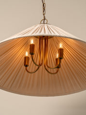 A pleated conical pendant light shade in a warm cream or off-white fabric, illuminated with a soft warm glow. It hangs from a dark brass chain and ceiling canopy against a plain, warm beige wall.