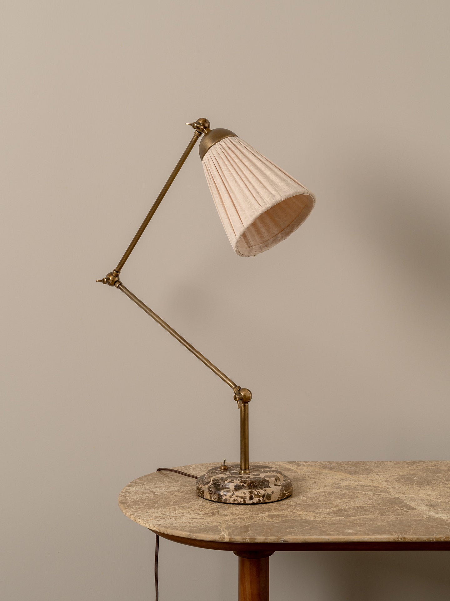 A mid-century modern adjustable desk lamp with a pleated beige shade, brass-colored metal arm, and a round stone base, shown on a stone-topped table against a neutral beige wall.