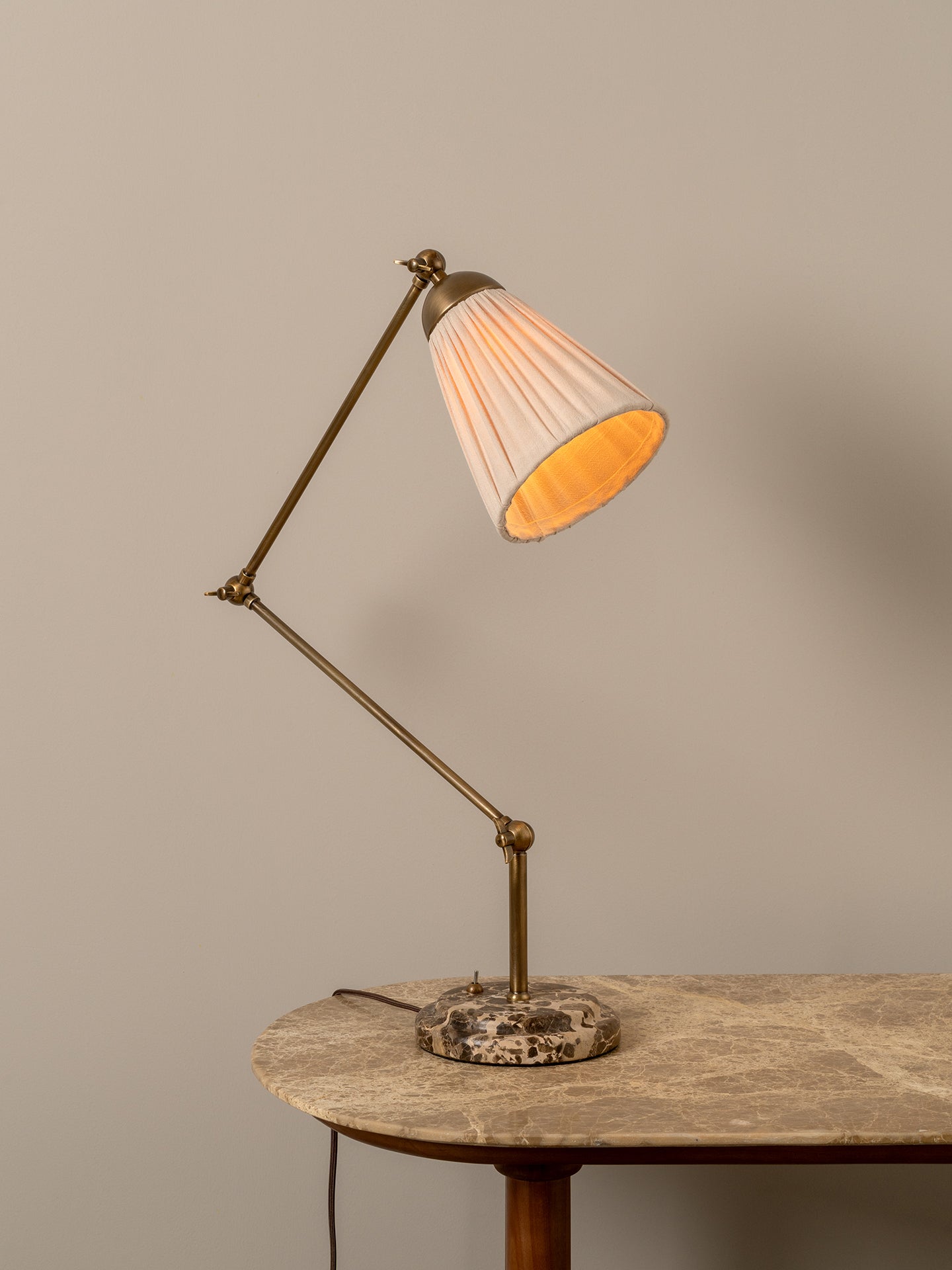 A brass adjustable desk lamp with a pleated off-white shade glowing warm yellow, set on a stone-topped table against a neutral beige wall.