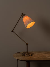 A classic adjustable desk lamp with a dark metal articulated arm, a pleated white shade glowing with warm orange light, and a distressed round base, shown on a rustic wooden table against a dark gray background.
