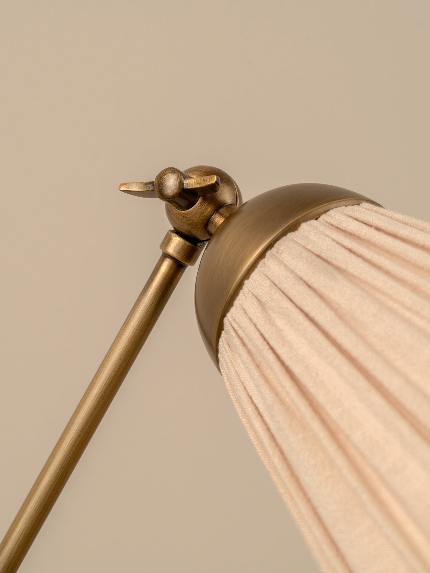 Close-up detail of a table lamp showing a brushed brass metal finial and pole with a pleated fabric shade in a soft blush pink, set against a solid warm beige background.