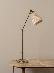 A mid-century modern adjustable desk lamp with a brass-colored metal stand and a pleated off-white shade, resting on a marble-topped table against a neutral beige wall.