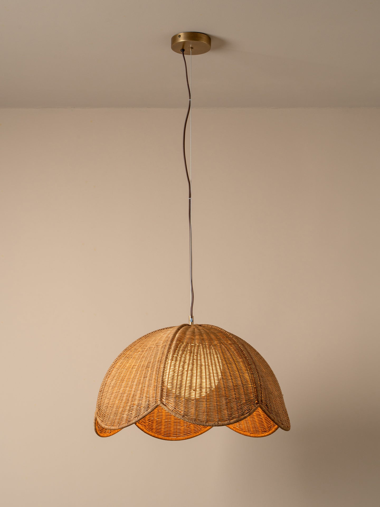 A warm-toned pendant light with a woven wicker shade hanging from a black cord and brass ceiling canopy against a plain beige wall.