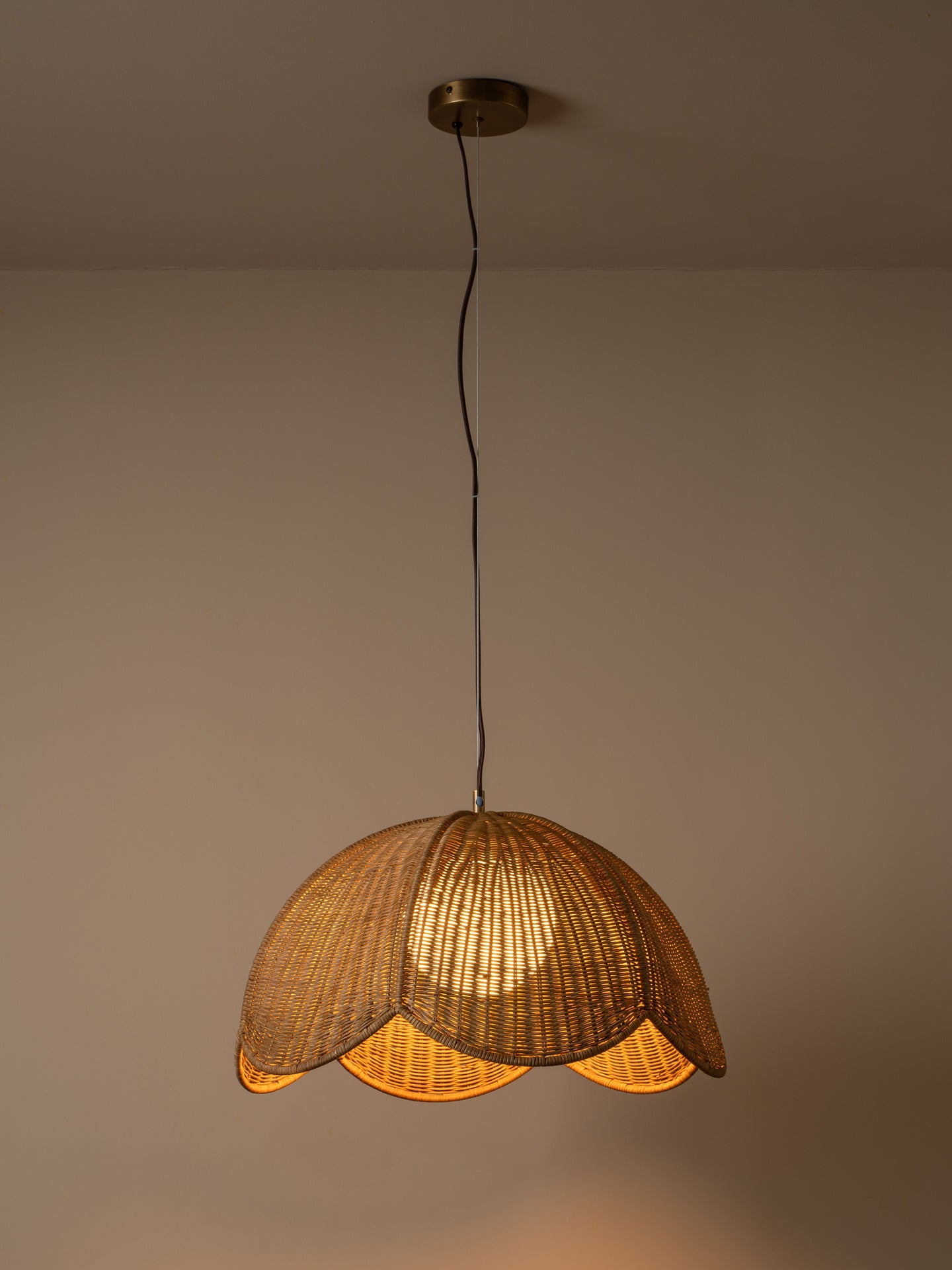 A warm-toned pendant light with a woven wicker shade hanging from a black cord and brass ceiling canopy against a plain beige wall.