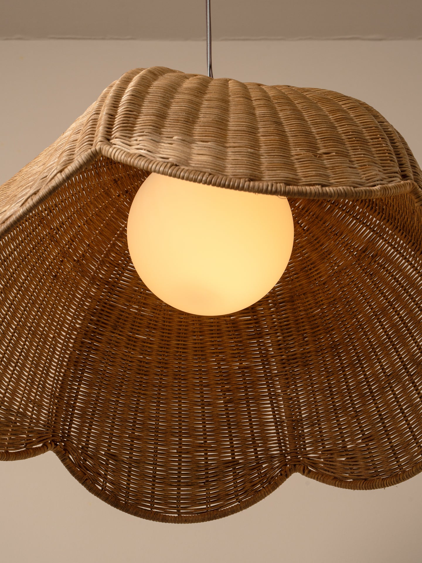 A close-up of a woven rattan pendant lamp shade with a glowing warm white globe bulb inside, set against a plain beige wall.