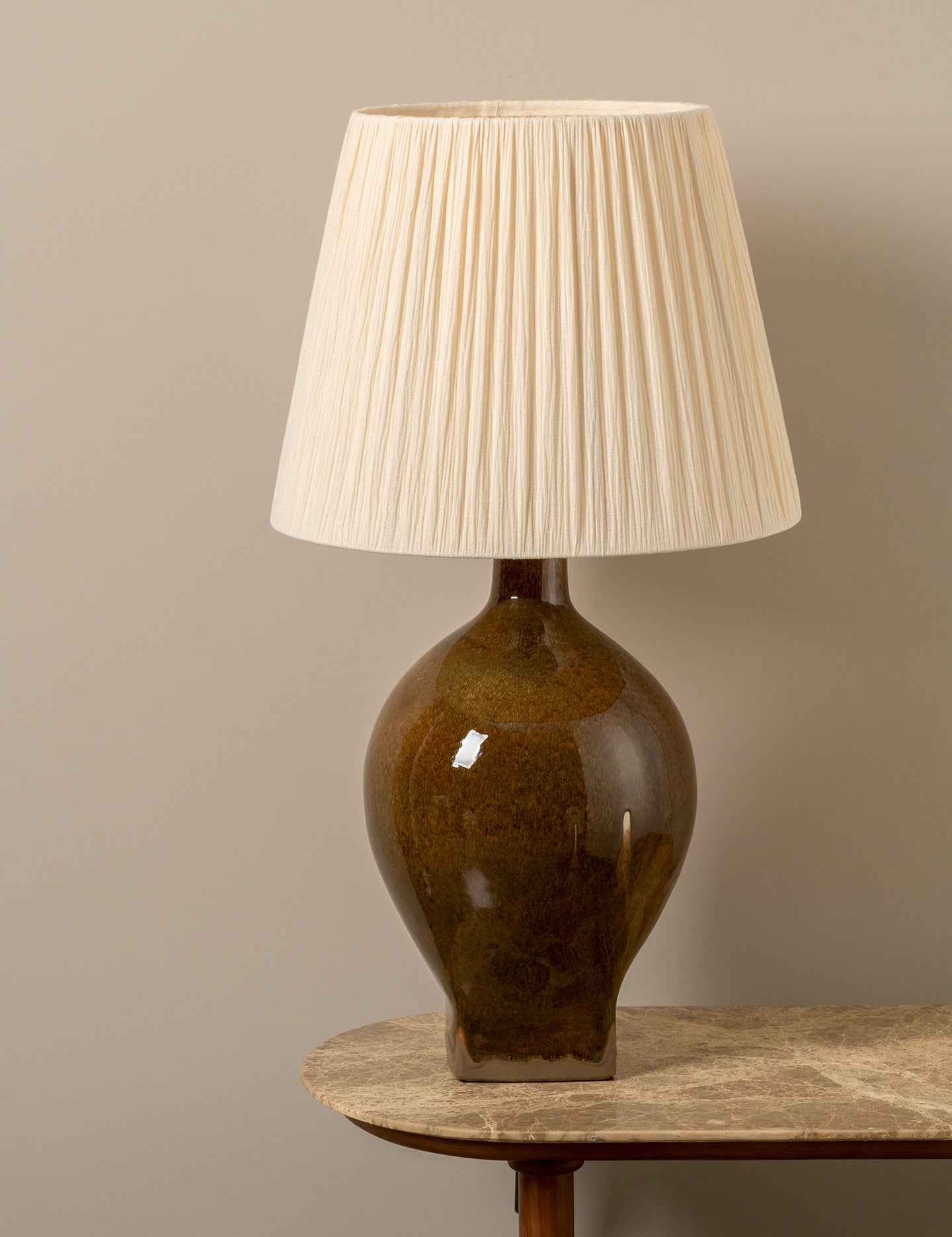 A table lamp with a pleated cream fabric shade and a glossy amber-brown ceramic base, shown on a stone-topped table against a neutral beige wall.