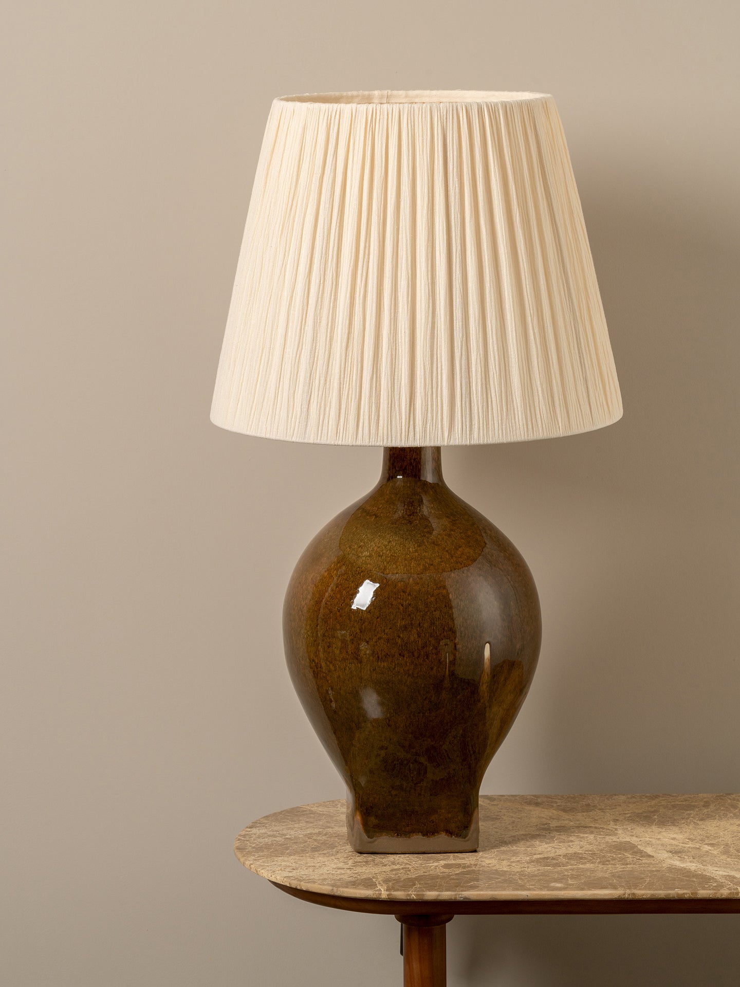 A table lamp with a pleated cream fabric shade and a glossy amber-brown ceramic base, shown on a stone-topped table against a neutral beige wall.