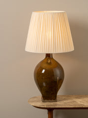 A table lamp with a glossy brown ceramic base and a pleated off-white fabric shade, illuminated and shown on a stone-topped table against a neutral beige wall.