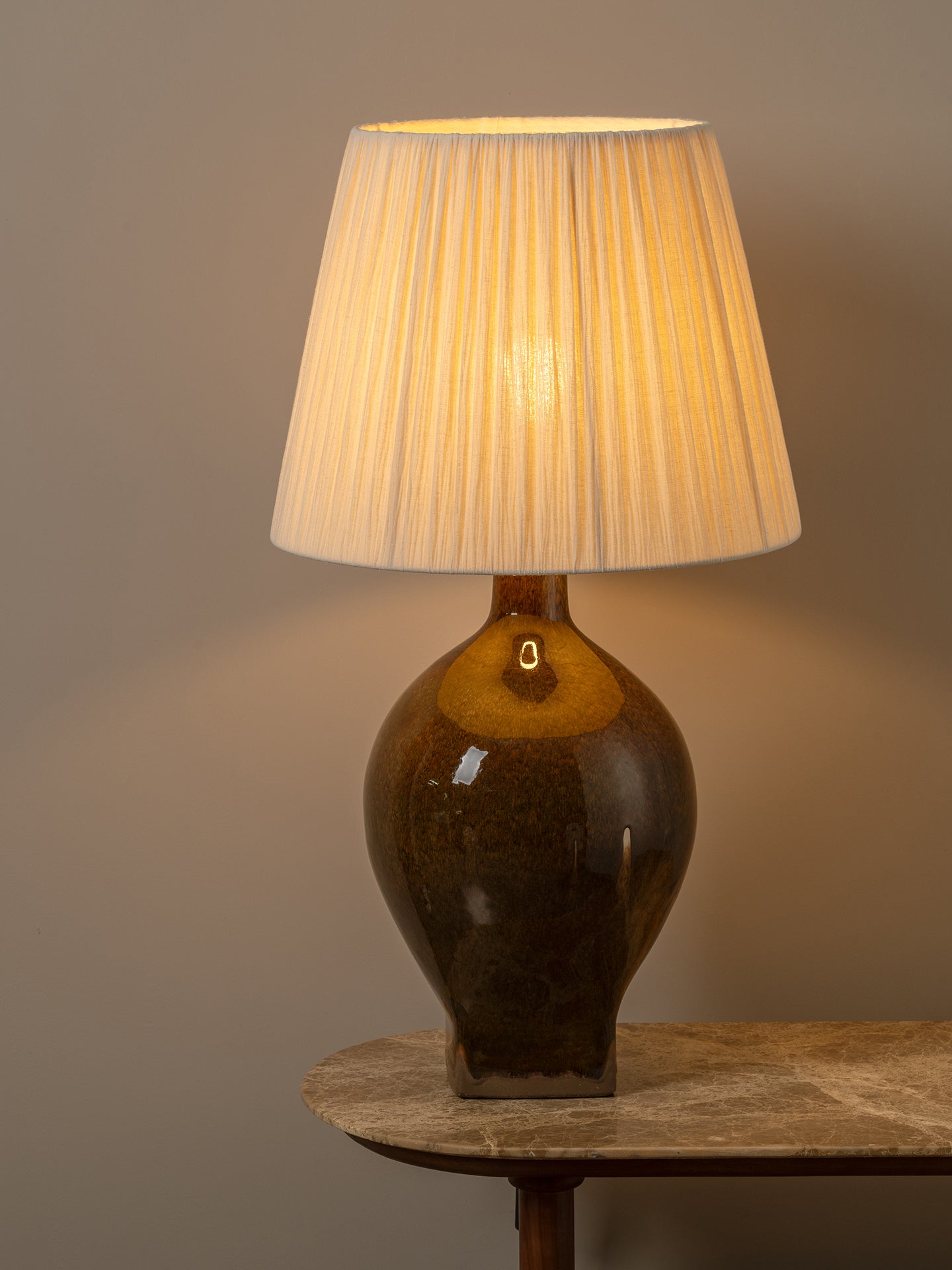 A table lamp with a pleated off-white fabric shade and a glossy dark brown ceramic base, illuminated to cast a warm glow against a neutral gray background.