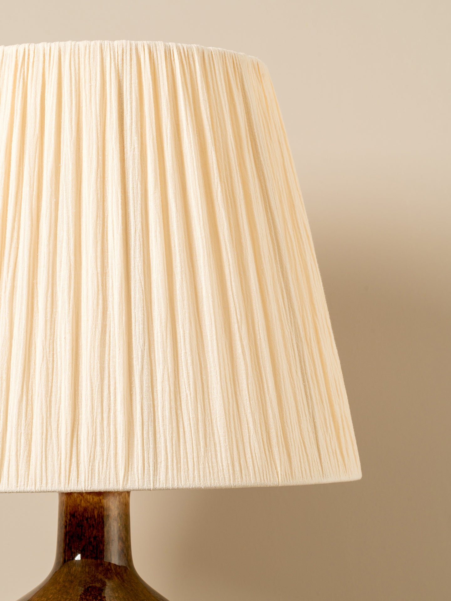 A close-up of a pleated fabric lampshade in a warm cream or off-white color, attached to a dark brown ceramic lamp base against a solid beige background.