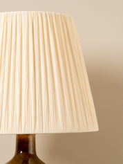 A close-up of a pleated fabric lampshade in a warm cream or off-white color, attached to a dark brown ceramic lamp base against a solid beige background.