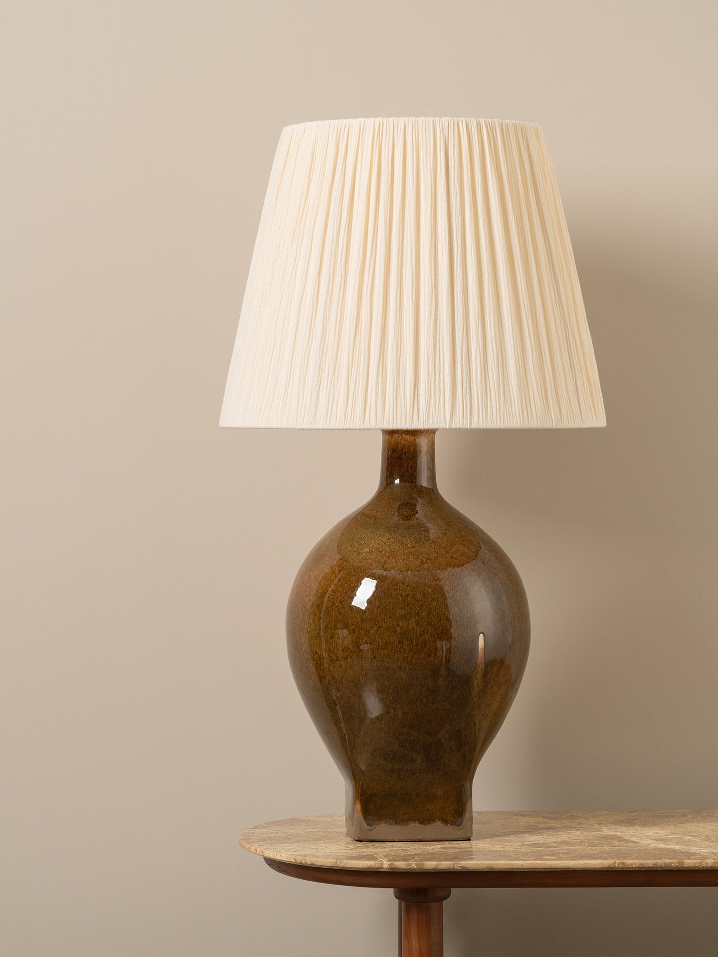 A table lamp with a glossy brown ceramic bulb and a cream pleated fabric shade, displayed on a wooden side table against a neutral beige wall.