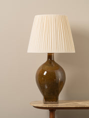 A table lamp with a glossy brown ceramic bulb and a cream pleated fabric shade, displayed on a wooden side table against a neutral beige wall.