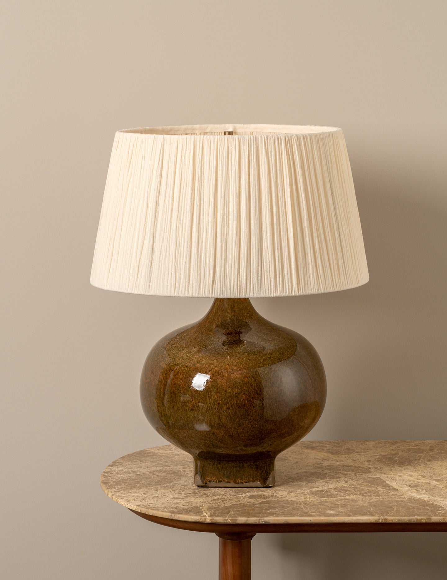 A table lamp with a glossy brown ceramic base and a pleated off-white fabric shade, displayed on a stone-topped side table against a neutral beige wall.