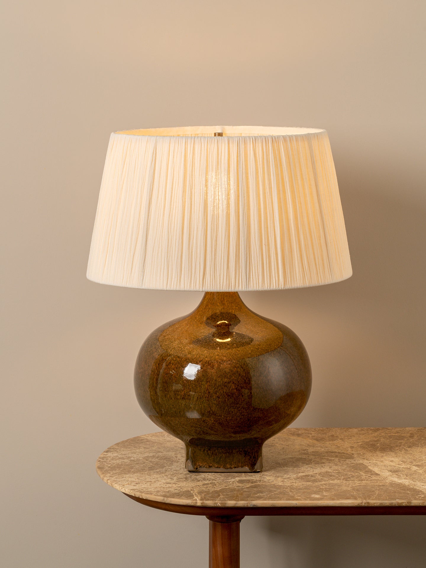 A table lamp with a glossy amber-brown ceramic base and a pleated off-white fabric shade, displayed on a stone-topped side table against a neutral beige wall.