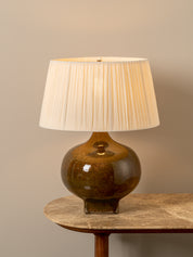A table lamp with a glossy amber-brown ceramic base and a pleated off-white fabric shade, displayed on a stone-topped side table against a neutral beige wall.