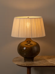 A table lamp with a pleated cream fabric shade and a glossy dark brown ceramic base, shown lit on a stone-topped table against a warm gray background.