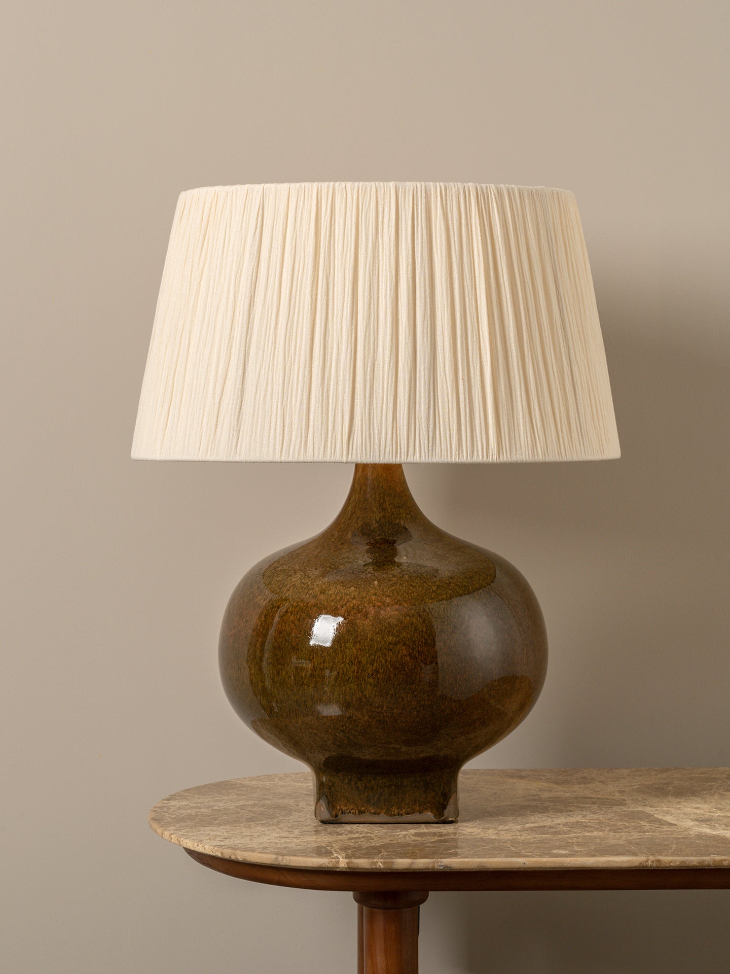 A table lamp with a glossy brown ceramic bulb and a pleated off-white fabric shade, displayed on a wooden table against a neutral beige background.