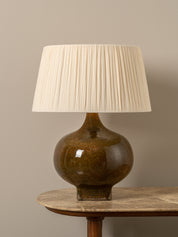 A table lamp with a glossy brown ceramic bulb and a pleated off-white fabric shade, displayed on a wooden table against a neutral beige background.