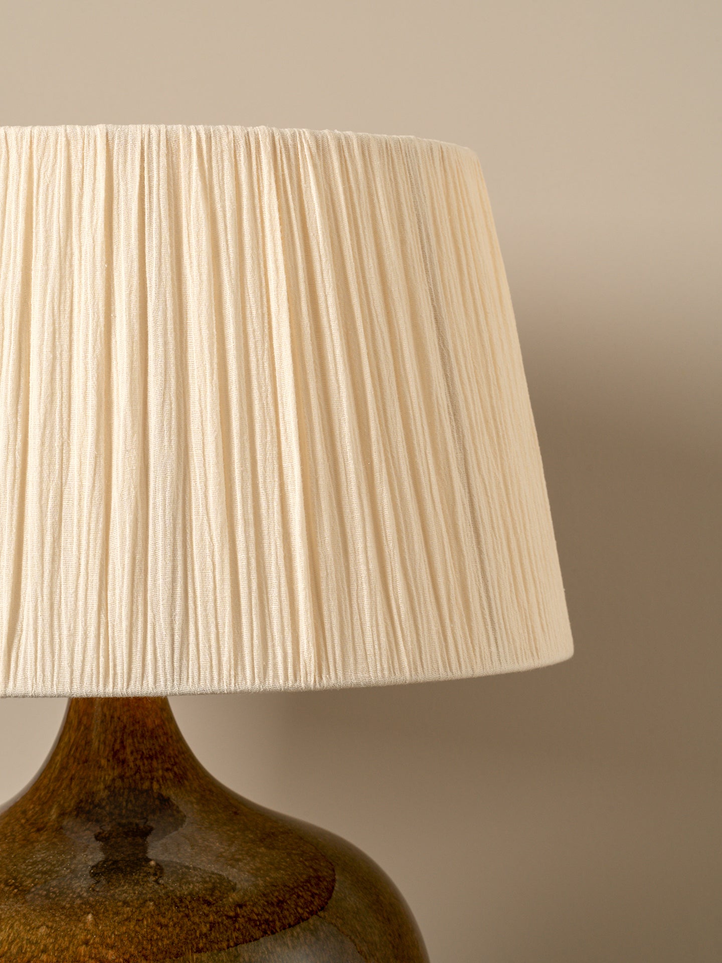 A close-up of a pleated fabric lampshade in a warm cream color, attached to a glossy amber-brown ceramic lamp base against a neutral beige background.