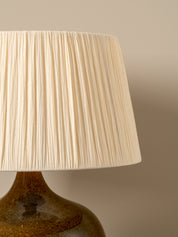 A close-up of a pleated fabric lampshade in a warm cream color, attached to a glossy amber-brown ceramic lamp base against a neutral beige background.