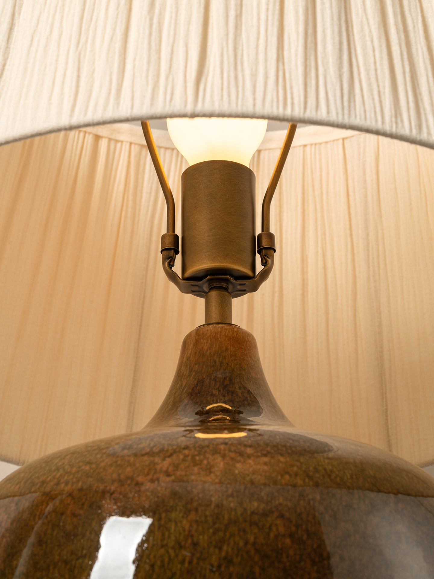 Close-up of a table lamp with a glossy brown ceramic base, brass metal socket, and a cream pleated fabric lampshade, with the light turned on.