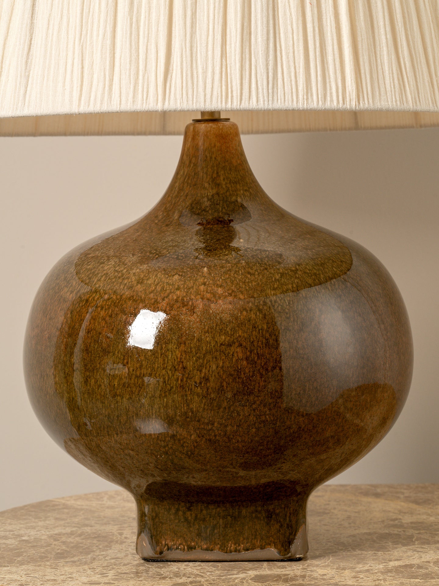 A ceramic table lamp with a rounded bulbous base in a glossy brown and amber glaze, topped with a cream pleated fabric shade, shown on a tabletop against a neutral beige background.