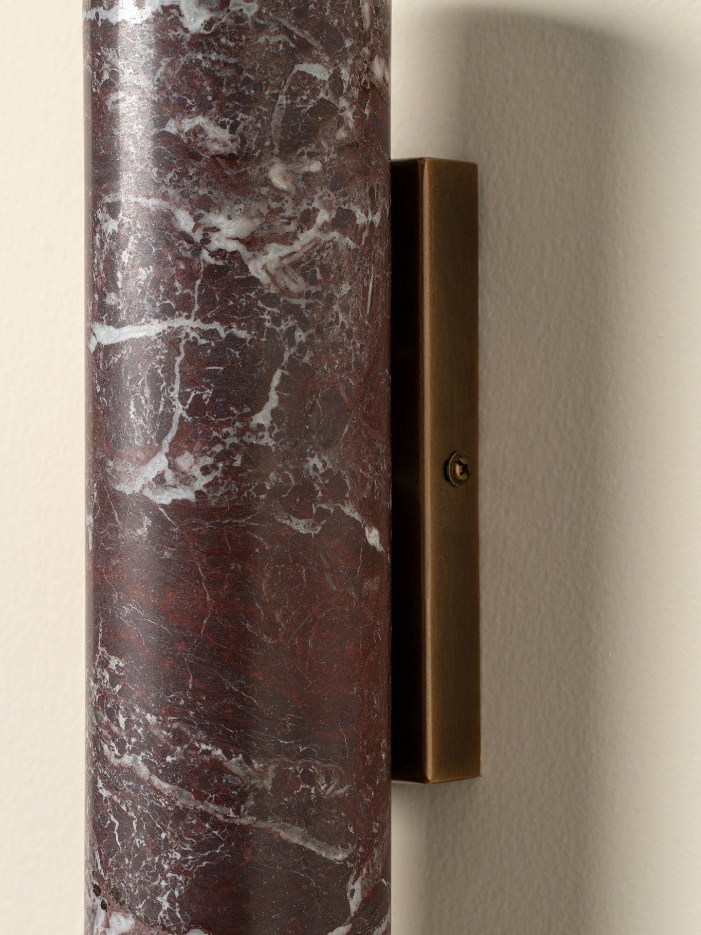 A close-up of a wall-mounted light fixture with a cylindrical dark red marble shade featuring white veining and a rectangular bronze metal backplate on a beige wall.