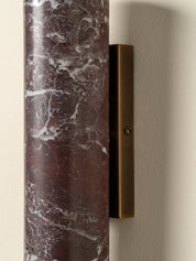 A close-up of a wall-mounted light fixture with a cylindrical dark red marble shade featuring white veining and a rectangular bronze metal backplate on a beige wall.