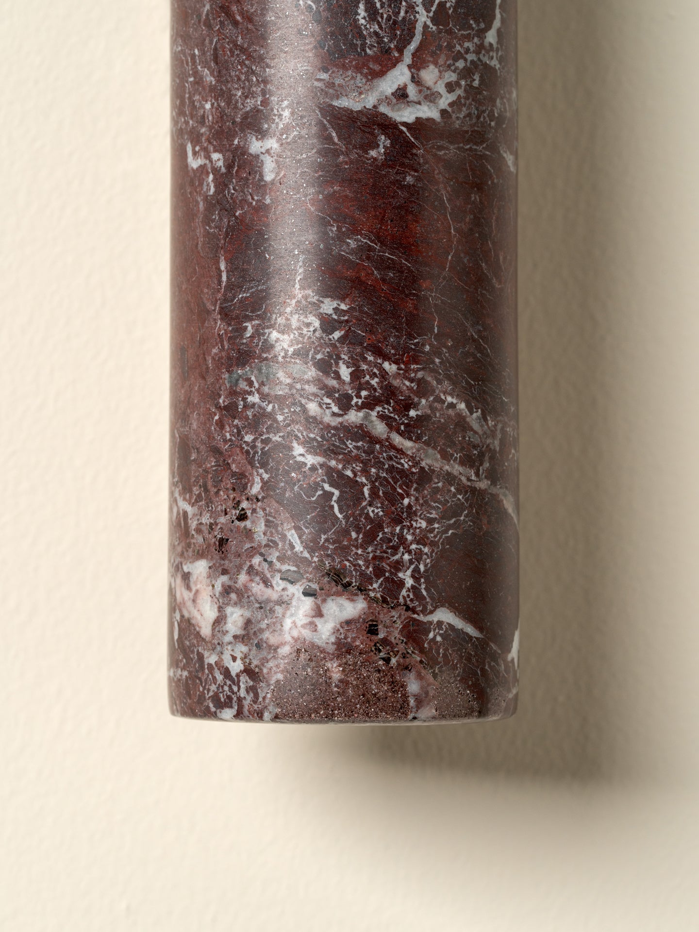 A close-up of a cylindrical wall sconce made from polished dark red marble with white and gray veining, mounted on a beige wall.