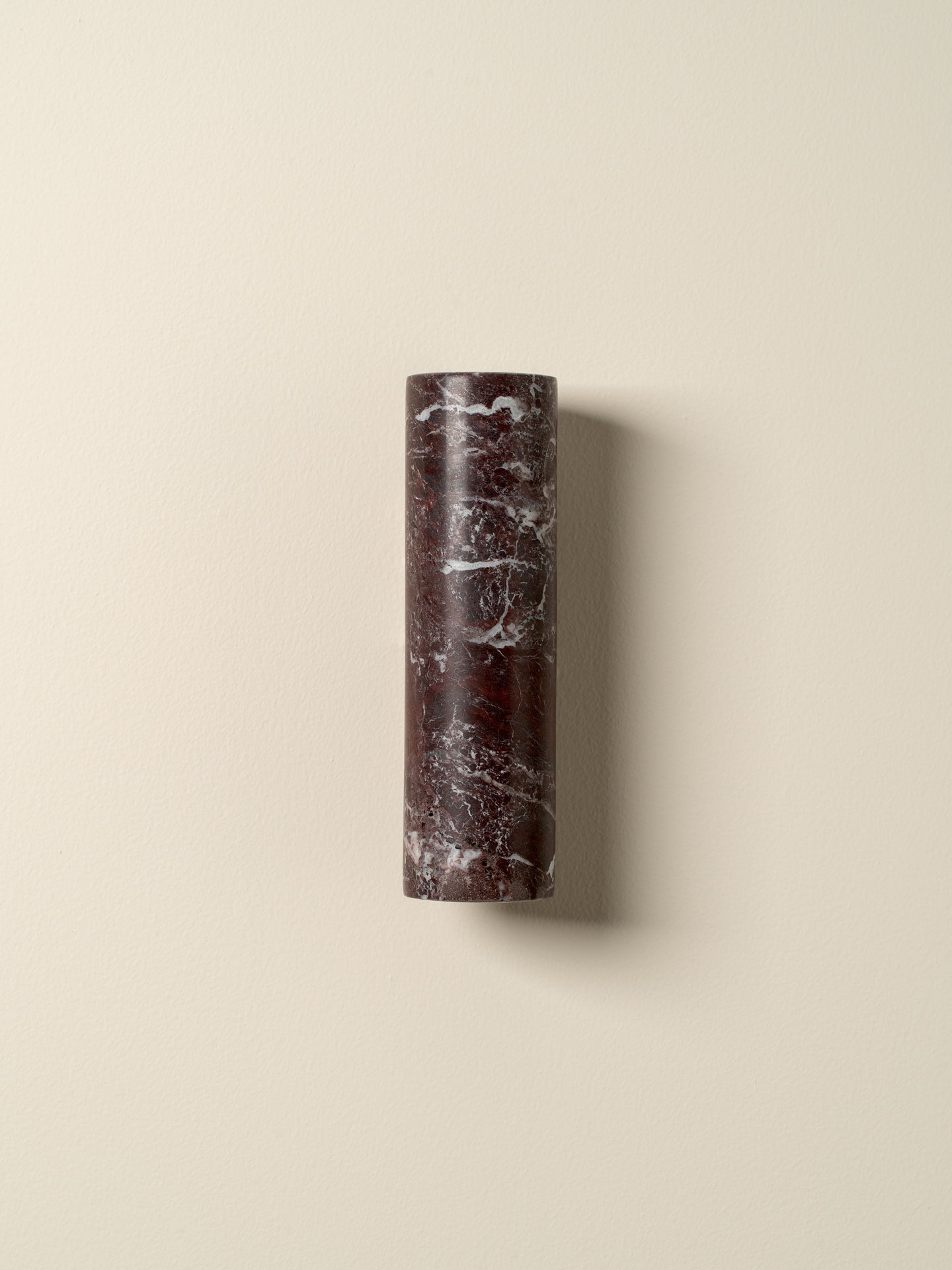 A cylindrical wall sconce made of dark reddish-brown marble with white veining, mounted on a warm beige wall.