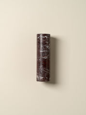 A cylindrical wall sconce made of dark reddish-brown marble with white veining, mounted on a warm beige wall.