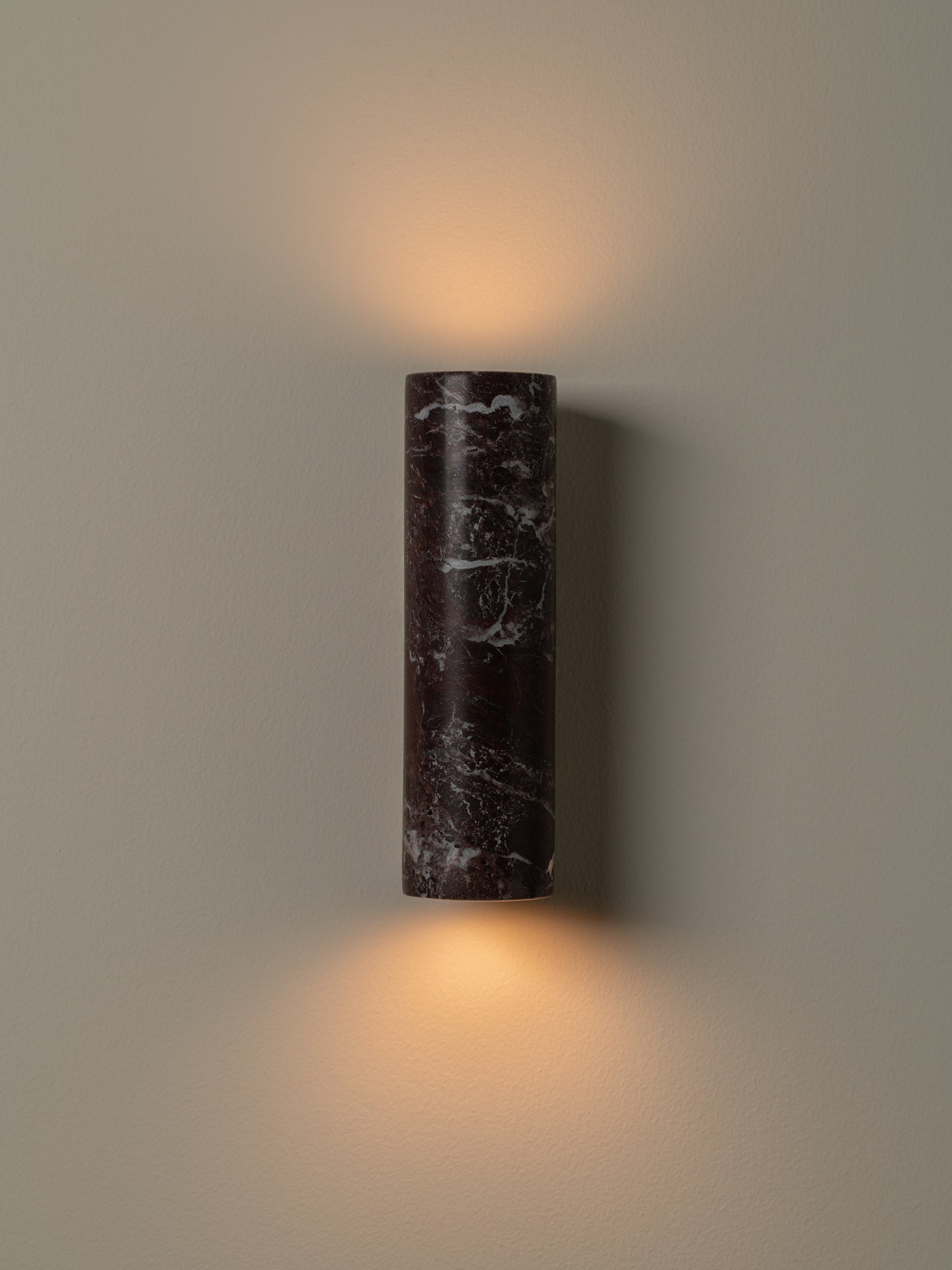 A cylindrical wall sconce made of dark marble with white veining, mounted on a neutral beige wall and casting a warm light upward and downward.