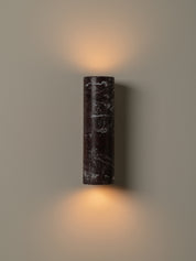 A cylindrical wall sconce made of dark marble with white veining, mounted on a neutral beige wall and casting a warm light upward and downward.