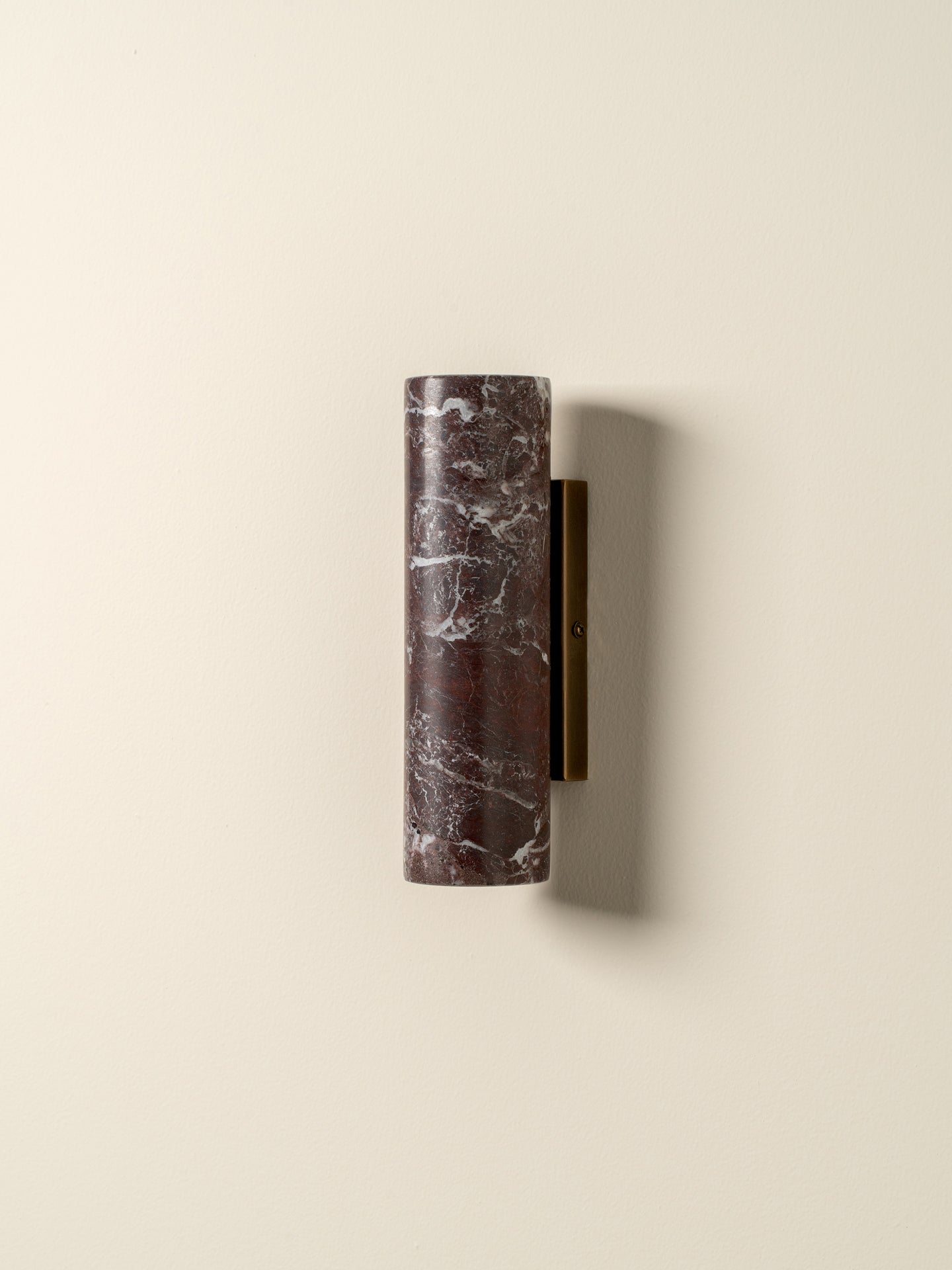 A modern cylindrical wall sconce featuring a dark reddish-brown marble shade with white veining and a matte brass metal bracket, mounted on a warm off-white wall.