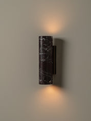 A modern cylindrical wall sconce with a dark marble-textured shade and a black metal mount, shown illuminated against a warm neutral wall.
