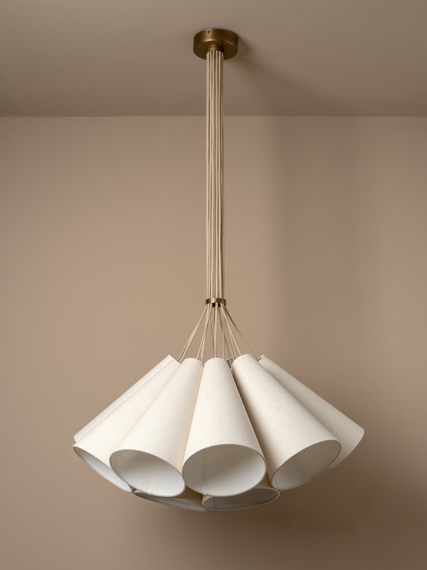 A modern multi-light cluster, pendant chandelier with five tapered off white linen shades fanned out from a central aged brass ceiling cup, suspended by oatmeal beige cables against a solid warm beige background.