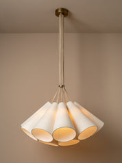 A modern multi-light cluster, pendant chandelier with five tapered off white linen shades fanned out from a central aged brass ceiling cup, suspended by oatmeal beige cables against a solid warm beige background.