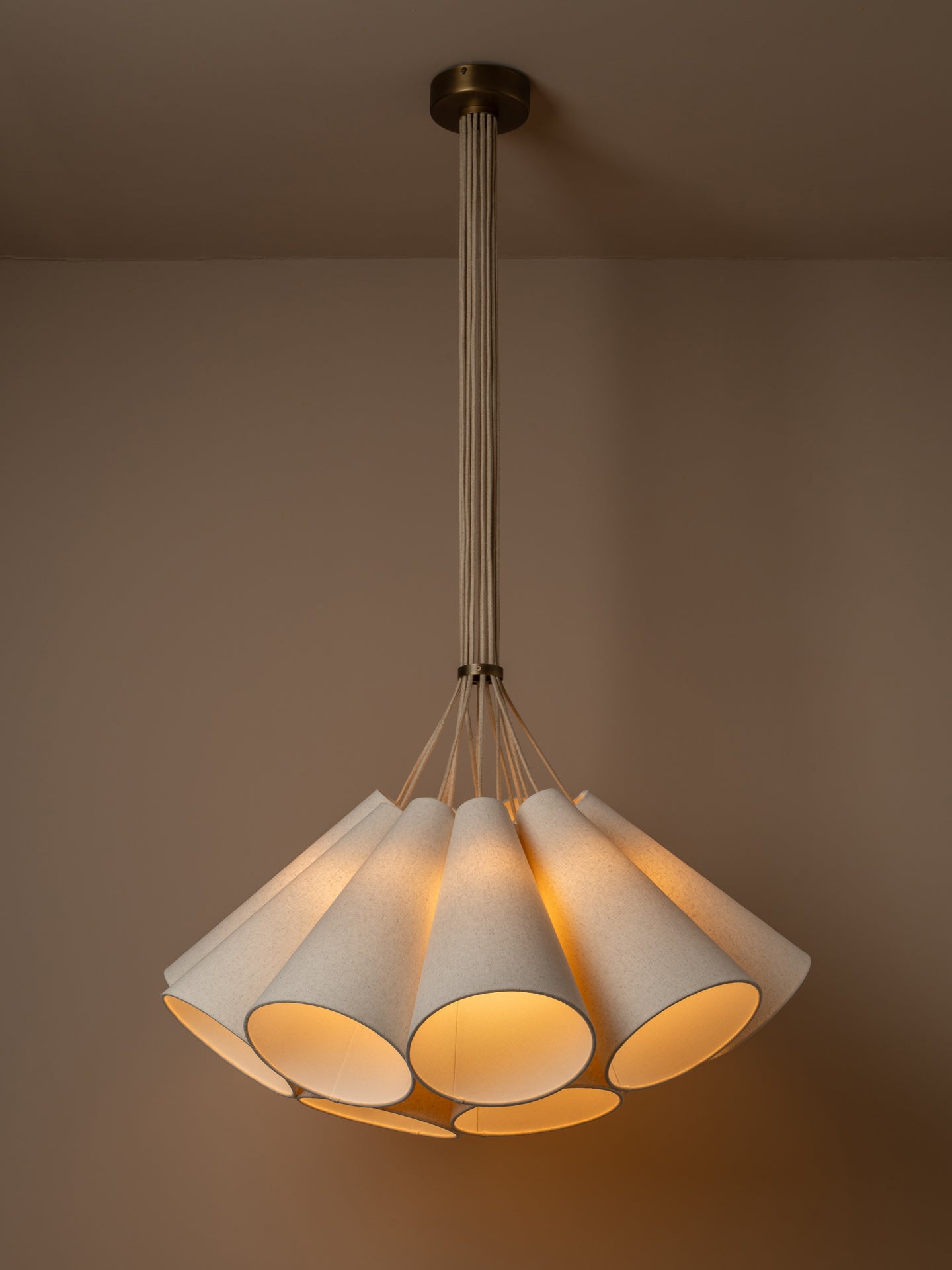 A modern multi-light cluster, pendant chandelier with five tapered off white linen shades fanned out from a central aged brass ceiling cup, suspended by oatmeal beige cables against a solid warm beige background.