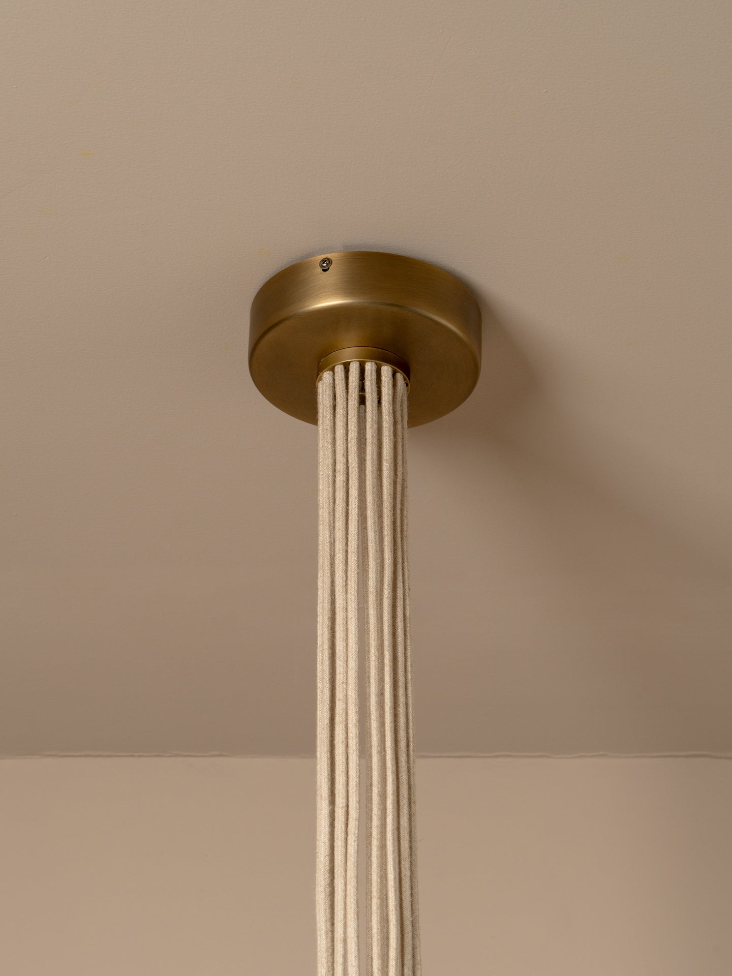 A modern multi-light cluster, pendant chandelier with five tapered off white linen shades fanned out from a central aged brass ceiling cup, suspended by oatmeal beige cables against a solid warm beige background.