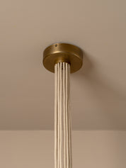A modern multi-light cluster, pendant chandelier with five tapered off white linen shades fanned out from a central aged brass ceiling cup, suspended by oatmeal beige cables against a solid warm beige background.
