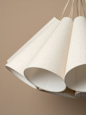 A modern multi-light cluster, pendant chandelier with five tapered off white linen shades fanned out from a central aged brass ceiling cup, suspended by oatmeal beige cables against a solid warm beige background.