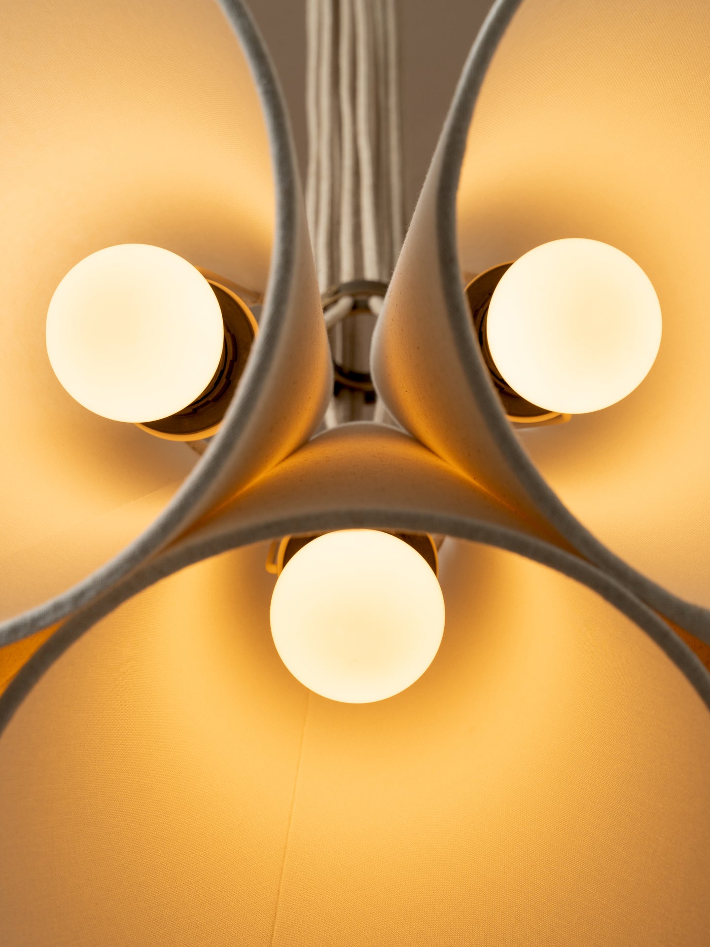 A modern multi-light cluster, pendant chandelier with five tapered off white linen shades fanned out from a central aged brass ceiling cup, suspended by oatmeal beige cables against a solid warm beige background.