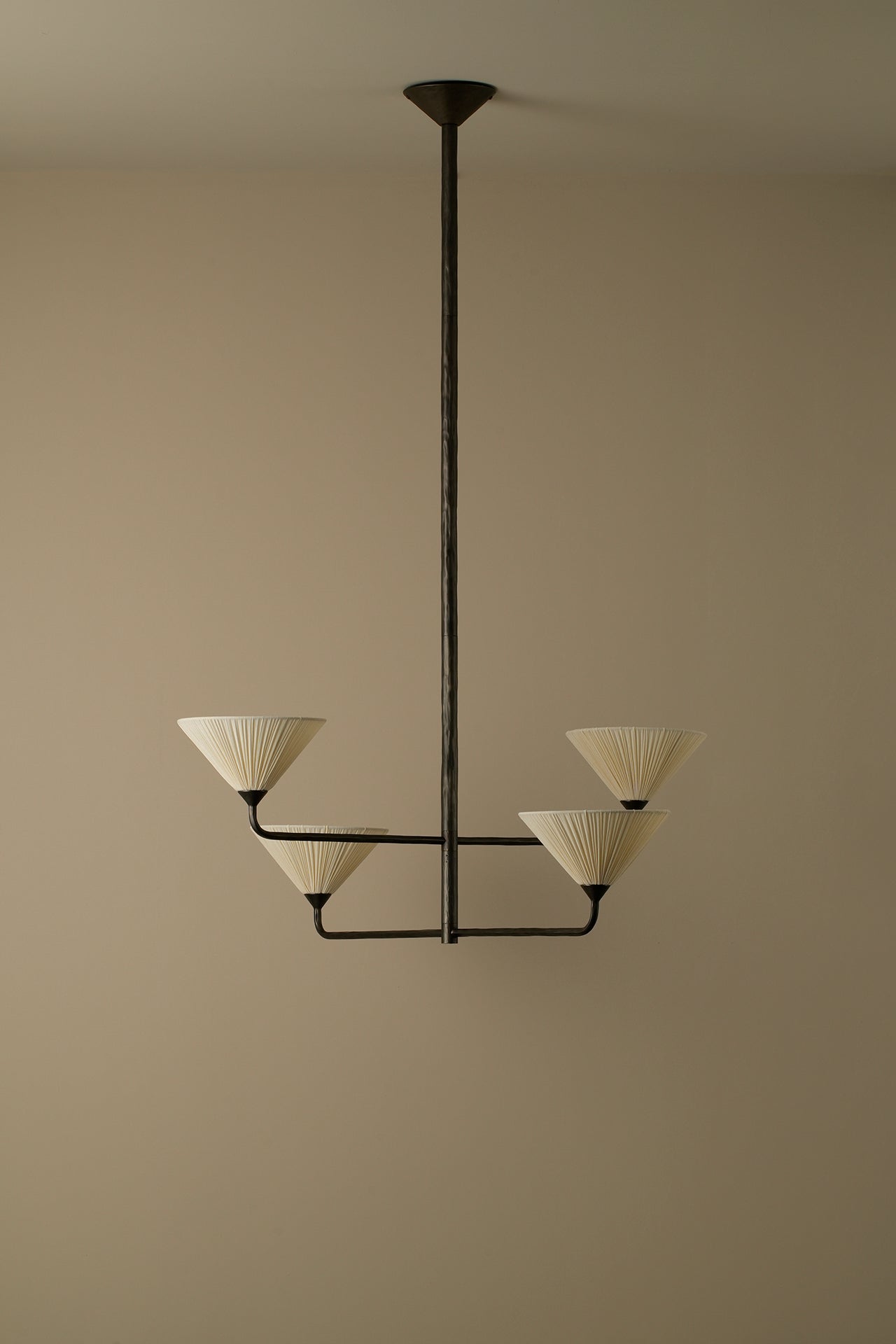 A modern multi-light chandelier with a dark metal frame and four cream pleated conical shades, suspended from a ceiling against a warm beige wall.